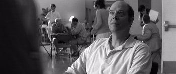Movie still from “Memento” (2000), directed by Christopher Nolan – Black and white photograph of a man sitting in front of a group of people; Close Up shot, Low angle