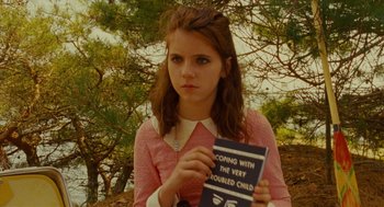 Movie still from “Moonrise Kingdom” (2012), directed by Wes Anderson – A young woman holding a book in front of a tree; Medium shot, High angle