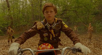 Movie still from “Moonrise Kingdom” (2012), directed by Wes Anderson – A boy scout riding a bike through the woods; Medium shot, Low angle