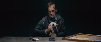 Movie still from “Nobody” (2021), directed by Ilya Naishuller – A man holding a kitten while sitting at a table; Medium shot, Overhead angle