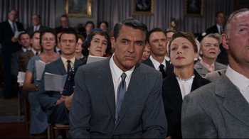 Movie still from “North by Northwest” (1959), directed by Alfred Hitchcock – A man in a suit and tie sitting in front of a group of people; Medium shot, Over the shoulder angle