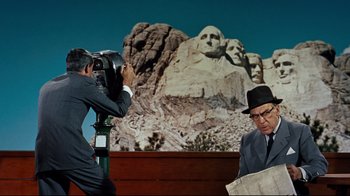 Movie still from “North by Northwest” (1959), directed by Alfred Hitchcock – A man in a suit and a hat standing next to a man in a suit and a hat; Wide shot, Low angle