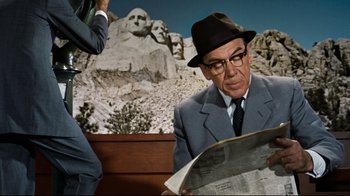 Movie still from “North by Northwest” (1959), directed by Alfred Hitchcock – An older man in a suit and hat is reading a newspaper; Medium shot, Over the shoulder angle