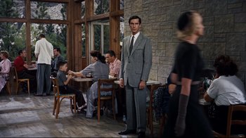 Movie still from “North by Northwest” (1959), directed by Alfred Hitchcock – A man in a suit standing in front of a group of people in a restaurant; Wide shot, Over the shoulder angle
