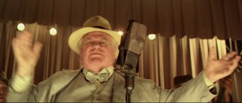 Movie still from “O Brother, Where Art Thou?” (2000), directed by Joel Coen – An older man wearing a hat and holding a microphone in front of him; Close Up shot, Low angle