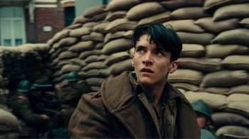 Movie still from “Dunkirk” (2017), directed by Christopher Nolan – A young man in a trench coat looking off into the distance in front of stacks of sandbags; Close Up shot, Low angle