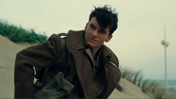 Movie still from “Dunkirk” (2017), directed by Christopher Nolan – A young man in a trench coat standing in the sand; Close Up shot, Low angle