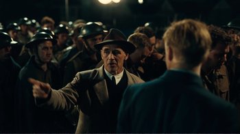 Movie still from “Dunkirk” (2017), directed by Christopher Nolan – An older man wearing a hat talking to a group of people; Medium shot, Over the shoulder angle