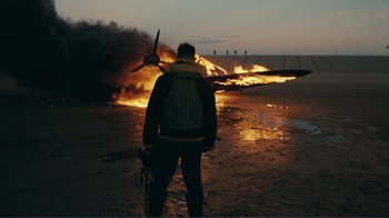 Movie still from “Dunkirk” (2017), directed by Christopher Nolan – A man standing in front of an airplane that is on fire; Wide shot, Over the shoulder angle