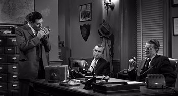 Movie still from “Psycho” (1960), directed by Alfred Hitchcock – A man sitting at a desk in front of two other men; Medium shot, Over the shoulder angle