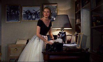 Movie still from “Rear Window” (1954), directed by Alfred Hitchcock – A woman in a black top and a white skirt is standing in front of a desk; Medium shot, Low angle