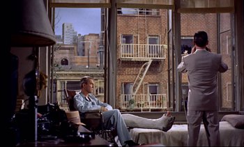 Movie still from “Rear Window” (1954), directed by Alfred Hitchcock – A man sitting in a chair in front of a large window; Wide shot, Over the shoulder angle