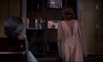 Movie still from “Rear Window” (1954), directed by Alfred Hitchcock – A woman in a pink dress standing in front of a building; Wide shot, Over the shoulder angle