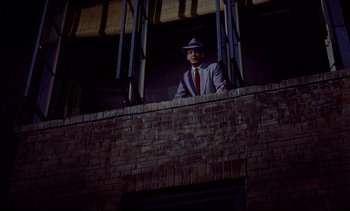 Movie still from “Rear Window” (1954), directed by Alfred Hitchcock – A man in a suit and hat standing on a balcony; Wide shot, Low angle