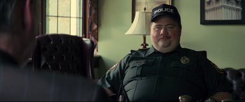 Movie still from “Richard Jewell” (2019), directed by Clint Eastwood – A man in a police uniform sitting in a chair in front of a lamp; Medium shot, Over the shoulder angle
