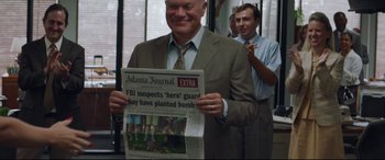 Movie still from “Richard Jewell” (2019), directed by Clint Eastwood – A man in a suit and tie holding up a newspaper; Medium shot, Over the shoulder angle