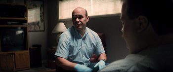 Movie still from “Richard Jewell” (2019), directed by Clint Eastwood – A bald man wearing a blue and white striped shirt and blue gloves; Medium shot, Over the shoulder angle