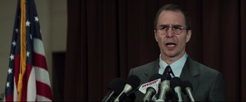 Movie still from “Richard Jewell” (2019), directed by Clint Eastwood – A man in a gray suit is speaking into microphones in front of a curtain; Close Up shot, Over the shoulder angle