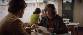 Movie still from “Rocketman” (2019), directed by Dexter Fletcher – A man sitting at a table in front of other people; Medium shot, Over the shoulder angle