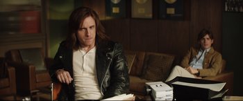 Movie still from “Rocketman” (2019), directed by Dexter Fletcher – A man sitting in a chair wearing a leather jacket; Medium shot, Over the shoulder angle