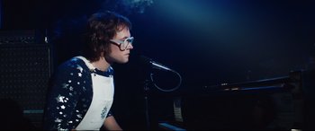 Movie still from “Rocketman” (2019), directed by Dexter Fletcher – A man with glasses is singing into a microphone; Medium shot, Low angle
