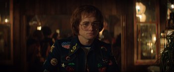 Movie still from “Rocketman” (2019), directed by Dexter Fletcher – A man in a jacket and glasses is standing in a room; Medium shot, Over the shoulder angle