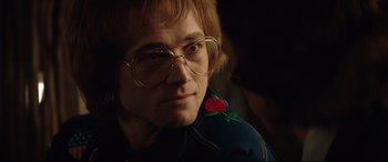 Movie still from “Rocketman” (2019), directed by Dexter Fletcher – A man with a rose on his shoulder wearing round glasses; Close Up shot, Over the shoulder angle