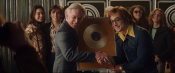Movie still from “Rocketman” (2019), directed by Dexter Fletcher – Two men shaking hands while holding a plaque; Medium shot, Over the shoulder angle