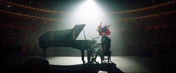 Movie still from “Rocketman” (2019), directed by Dexter Fletcher – A person in a colorful costume sitting at a piano; Wide shot, High angle
