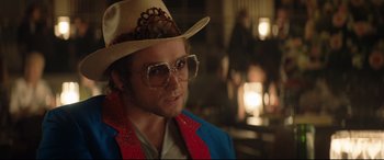 Movie still from “Rocketman” (2019), directed by Dexter Fletcher – A man wearing a hat and glasses in a room; Close Up shot, Over the shoulder angle