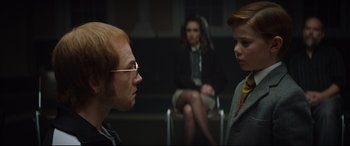 Movie still from “Rocketman” (2019), directed by Dexter Fletcher – A man and a woman are sitting in front of another man; Medium shot, Over the shoulder angle