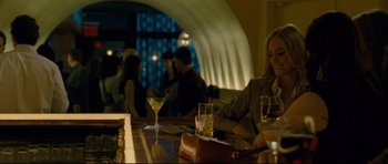 Movie still from “Shame” (2011), directed by Steve McQueen – A woman sitting at a table with a drink in a glass; Medium shot, Over the shoulder angle