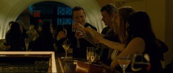Movie still from “Shame” (2011), directed by Steve McQueen – A group of people sitting at a table with drinks; Medium shot, Over the shoulder angle