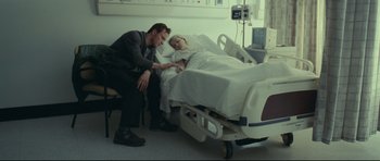 Movie still from “Shame” (2011), directed by Steve McQueen – A man sitting next to a woman laying in a hospital bed; Medium shot, High angle