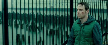 Movie still from “Shame” (2011), directed by Steve McQueen – A man standing in front of a fence with a crowd of people behind it; Medium shot, Over the shoulder angle