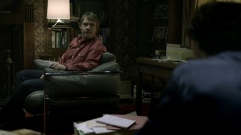 Movie still from “Sherlock” (2010), created by Steven Moffat – A man sitting in a chair in a living room; Wide shot, Over the shoulder angle