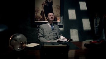 Movie still from “Sherlock” (2010), created by Steven Moffat – A man sitting at a table in front of a painting; Medium shot, Low angle