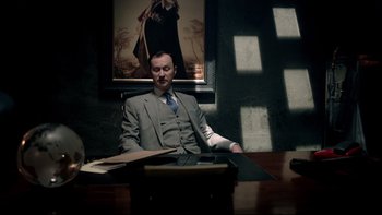 Movie still from “Sherlock” (2010), created by Steven Moffat – A man sitting at a desk in a suit and tie; Medium shot, Low angle
