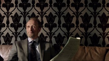 Movie still from “Sherlock” (2010), created by Steven Moffat – A man sitting in front of a wall holding a piece of paper; Close Up shot, Over the shoulder angle