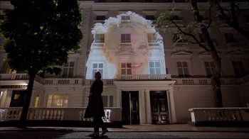 Movie still from “Sherlock” (2010), created by Steven Moffat – A woman is standing in front of a building with a picture on it; Wide shot, Low angle