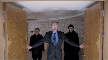 Movie still from “Sherlock” (2010), created by Steven Moffat – A man in a suit and tie standing between two men; Medium shot, Over the shoulder angle