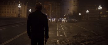Movie still from “Spectre” (2015), directed by Sam Mendes – A man holding a gun while standing on the side of a road; Wide shot, Over the shoulder angle
