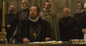 Movie still from “Elizabeth: The Golden Age” (2007), directed by Shekhar Kapur – A man sitting at a table in front of other men; Medium shot, Over the shoulder angle