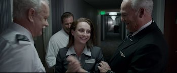 Movie still from “Sully” (2016), directed by Clint Eastwood – A woman in a business suit smiling for the camera; Medium shot, Over the shoulder angle