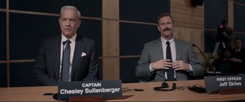 Movie still from “Sully” (2016), directed by Clint Eastwood – Two men sitting at a table in front of a sign; Medium shot, Over the shoulder angle