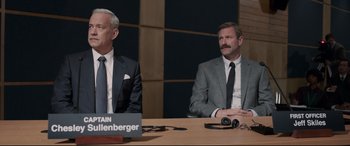 Movie still from “Sully” (2016), directed by Clint Eastwood – Two men sitting at a table in front of a microphone; Medium shot, Over the shoulder angle