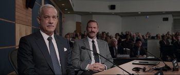 Movie still from “Sully” (2016), directed by Clint Eastwood – Two men in suits sitting at a podium in front of an audience; Medium shot, Over the shoulder angle