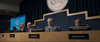 Movie still from “Sully” (2016), directed by Clint Eastwood – A man in a suit and tie sitting at a desk in front of an american flag; Medium shot, Over the shoulder angle