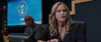 Movie still from “Sully” (2016), directed by Clint Eastwood – A woman with long blonde hair sitting in front of a microphone in front of a man; Medium shot, Over the shoulder angle