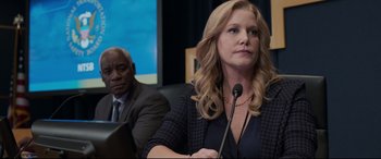 Movie still from “Sully” (2016), directed by Clint Eastwood – A woman is speaking at a microphone in front of a man; Close Up shot, Over the shoulder angle